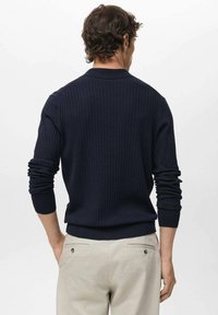Man wearing a navy ribbed sweater and beige pants, standing with hands in pockets, facing away against a plain white background.
