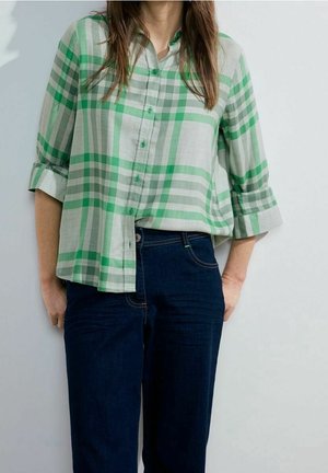 Femme portant une chemise à carreaux verte et blanche avec des boutons, manches retroussées, et un jean bleu foncé, debout contre un mur uni.
