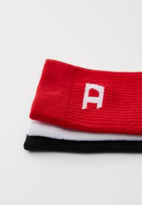 Trois bracelets colorés : rouge en haut avec un "A" blanc, suivi de blanc et noir, tous fabriqués en tissu doux et texturé.