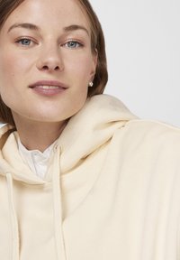 Joven con ojos azules que lleva pendientes de perlas y una sudadera con capucha crema sobre una camisa blanca abotonada, sonriendo suavemente.