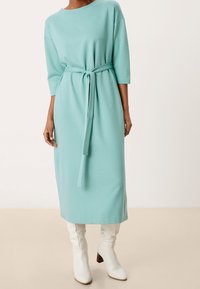 Un lungo vestito verde menta con una vestibilità morbida, maniche 3/4 e una cintura legata in vita. Abbinato a stivali bianchi con tacco al ginocchio.