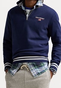 Navy blue zip-up sweatshirt with white striped cuffs, featuring "POLO SPORT" logo. Layered over a plaid shirt. Gray sweatpants visible below.