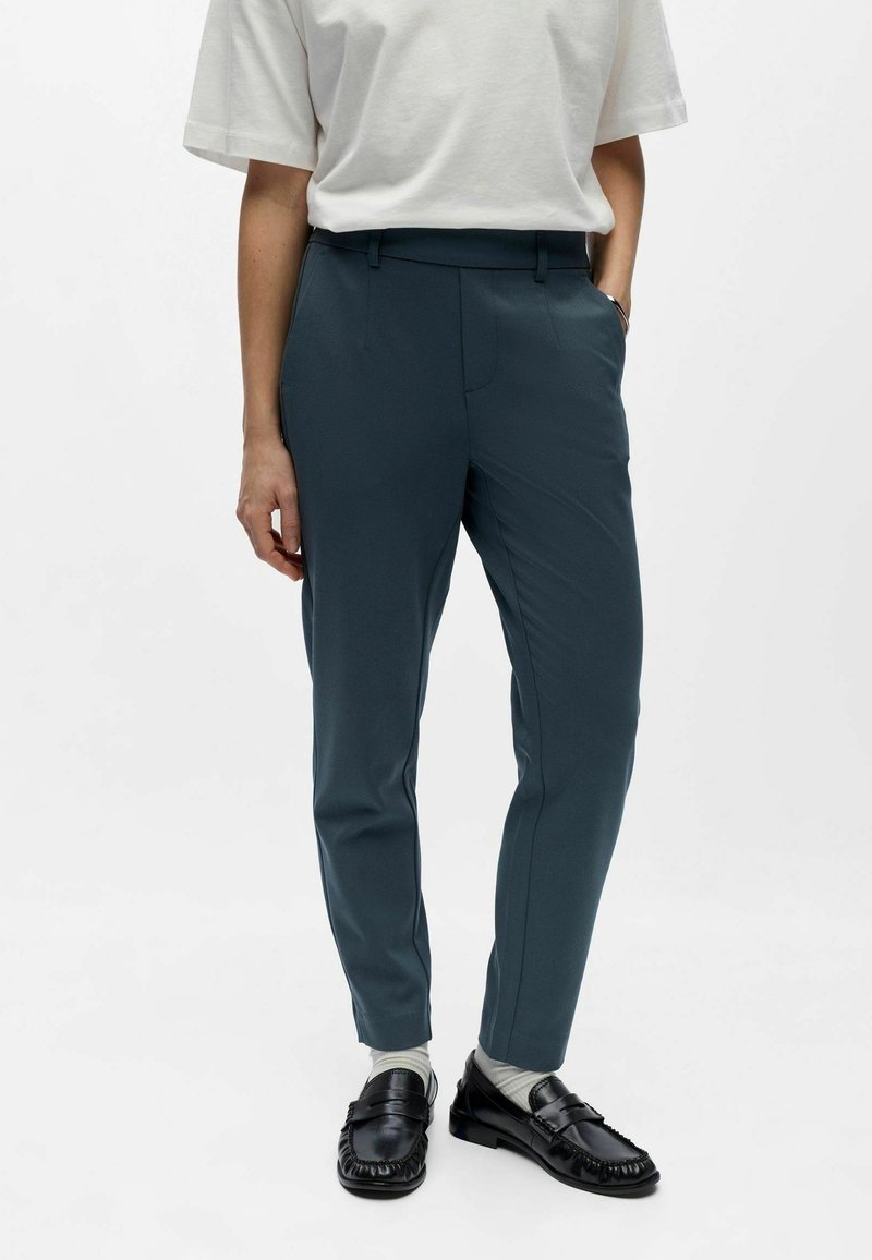 Personne portant un pantalon slim bleu sarcelle, un t-shirt blanc, des chaussettes blanches et des mocassins noirs, debout avec une main dans la poche.
