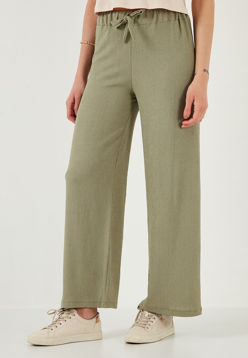 Licht olijfgroene, wijdvallende broek van een textuurstof, met een elastische tailleband en verstelbaar trekkoord; gecombineerd met beige sneakers.