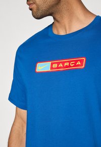 Μπλε βαμβακερό μπλουζάκι με στρογγυλή λαιμόκοψη, με πολύχρ�ωμο ορθογώνιο λογότυπο που περιλαμβάνει το κείμενο "BARÇA" και το λογότυπο της Nike.