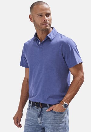 Polohemd aus leichtem lila Baumwollstoff, mit kurzen Ärmeln und klassischem Kragen. Kombiniert mit ausgebleichten blauen Jeans und einem braunen Gürtel, akzentuiert durch eine Uhr.
