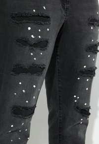 Zwarte verslete jeans met meerdere scheuren en rafelige randen, met witte spetteraccenten door de stof.