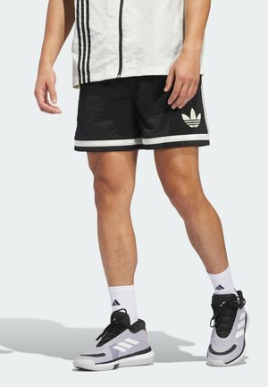 Beine einer Person, die schwarze Adidas-Shorts mit weißer Einfassung, weiße Adidas-Socken, graue und schwarze Adidas-Sneaker und ein weißes Oberteil mit schwarzen Streifen trägt.