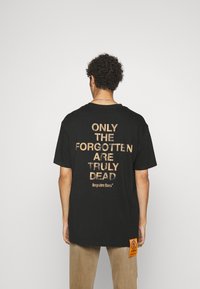 T-shirt en coton noir à manches courtes, avec un texte doré au dos : « SEULS LES OUBLIÉS SONT VRAIMENT MORTS. » Comprend une étiquette sur le côté.