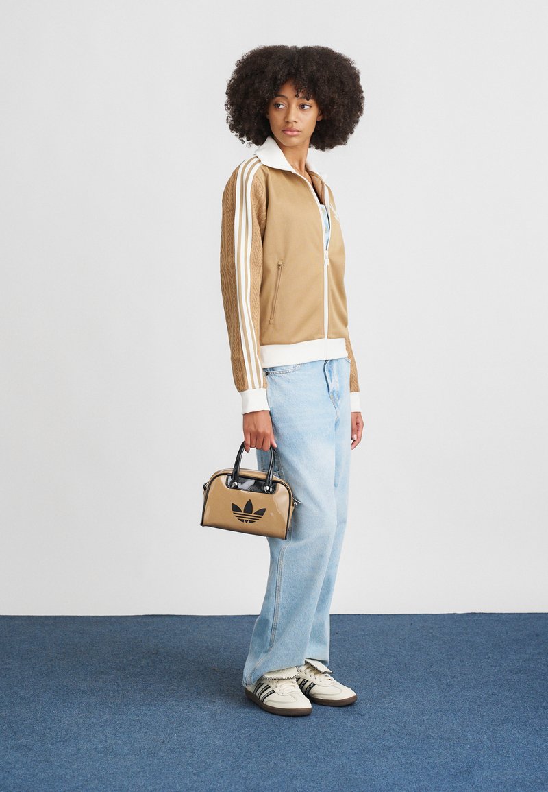 Braune Zip-Jacke mit weißen Streifen, hellblaue Jeans und eine kleine tanfarbene Handtasche mit schwarzem Logo, kombiniert mit weißen Turnschuhen.