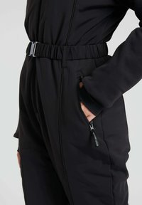 Schwarzer gepolsterter Jumpsuit mit tailliertem Bund, Reißverschlusstaschen und langen Ärmeln. Glatte Textur und figurbetontes Design.