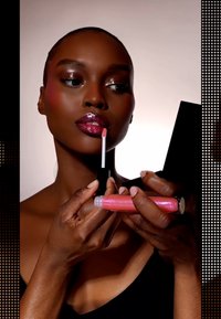 Lip gloss in a clear tube, with a pink, shimmery sheen. Model applies it with a wand, holding a black compact mirror. Smooth texture.