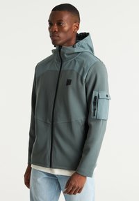 Capuchonjack in donkergroen en lichtteal, met een rits aan de voorkant, twee zijzakken en een mouwzak met hardware accenten. Gladde textuur.