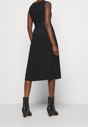 Nainen, joka on pukeutunut hihattomaan mustaan midimekkoon, jossa on laskokset, ja mustiin nilkkasaappaisiin, seisoo selin tasaisen valkoisen taustan edessä.