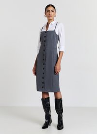 Graues Kleid mit Knopfleiste vorne, figurbetontem Schnitt, Trägern und seitlichen Taschen, kombiniert mit schwarzen kniehohen Stiefeln.