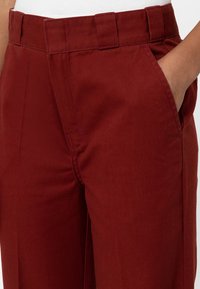 Burgundy byxor med rak skärning, sidofickor och en platt midja. Texturen är slät, med synliga sömnadsdetaljer.