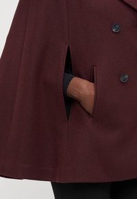 Burgundy kappa med en strukturerad design, som har två sidofickor och stora knappar. Materialet verkar ha en texturerad och ull-liknande yta.