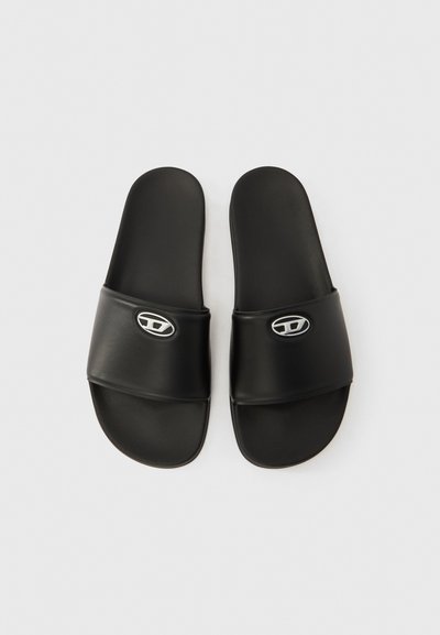 Sandales à enfiler noires en matière synthétique lisse, avec une large bride et un accent logo argenté sur le dessus. Semelle extérieure plate et texturée.