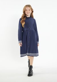 Marineblaue Kleid mit einem Volant-Ausschnitt, knöpfbarem Vorderteil und einem weißen Zickzacksaum. Kombiniert mit schwarzen Stiefeletten. Glattes Material, lange Ärmel.