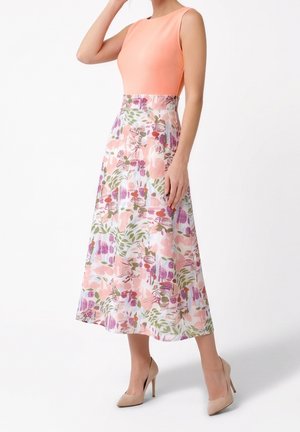 Femme portant un top pêche sans manches avec une jupe midi fleurie taille haute et des talons hauts beige, debout sur fond blanc.