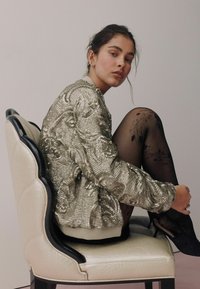 Metallic sequined bomber jacket with floral texture, cream ribbed collar, worn with sheer black tights featuring floral patterns.