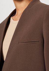 Blazer marrón con solapa chal, tela texturizada, un bolsillo en el pecho y combinado con una camisa beige clara.