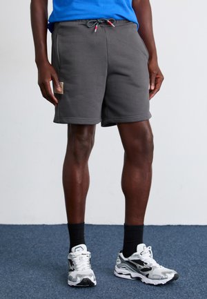 Shorts gris avec une taille élastique et un cordon de serrage, dotés d'une poche latérale. Assortis à des chaussettes noires et des chaussures de sport blanches.