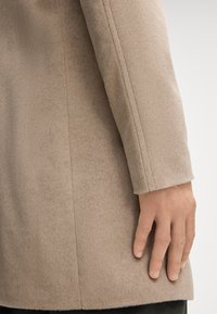 Beige wollen jas met een zachte textuur, voorzien van een rechte snit, zichtbare naden en een afgeronde mouwzoom. Hand die voorzichtig op de stof rust.