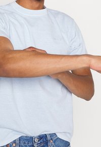 Světle modré bavlněné tričko s kulatým výstřihem a malým vystouplým logem. Ruka modelu je ohnutá, což ukazuje texturu látky a střih.