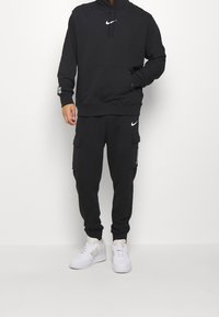Svart Nike-hoodie med en framficka, som har en huva och vitt logo. Kombinerad med svarta cargobyxor och vita sneakers.