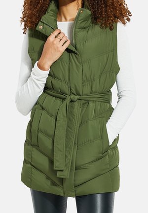 Nainen pukeutuneena hiukseton oliivinvihreään toppaliiviin, jossa on vyö vyötäröllä ja korkea kaulus, valkoiseen pitkähihainen paitaan ja mustiin housuihin, käsi taskussa.