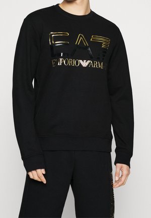 Person trägt einen schwarzen Pullover mit langen Ärmeln und glänzendem gold-schwarzem geprägtem EA7 Emporio Armani-Logo auf der Brust.