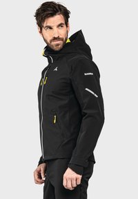 Schwarze Softshelljacke mit Kapuze, ausgestattet mit gelben Akzenten, Reißverschlusstaschen und reflektierenden Details an den Ärmeln. Hergestellt aus strukturiertem Material.