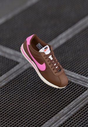 Brązowy sneaker Nike z różowym logo i białą podeszwą, wyświetlany na metalowej siatce.