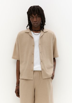 Chemise beige à manches courtes et boutons en tissu texturé, portée sur un t-shirt blanc. Associée avec un short beige assorti et des colliers superposés.