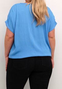 Personne aux longs cheveux blonds portant un haut bleu à manches courtes ample et un pantalon noir ajusté, vue de dos contre un mur uni.