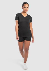 Czarny T-shirt z dekoltem w serek wykonany z elastycznej tkaniny, zestawiony z czarnymi szortami. Model nosi białe sneakersy, stoi na tle jasnoszarym.