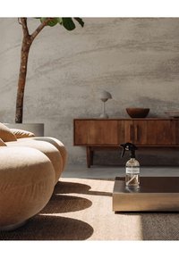 En genomskinlig sprayflaska med en svart trigger- sprejtop, märkt med text, placerad på ett metallbord över en vävd matta, bredvid en rund beige soffa.