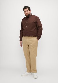 Brun langermet skjorte med knapper, beige bukser og hvite joggesko. Skjorten har klassisk krage og knapper, smal passform. Enkel tekstur.