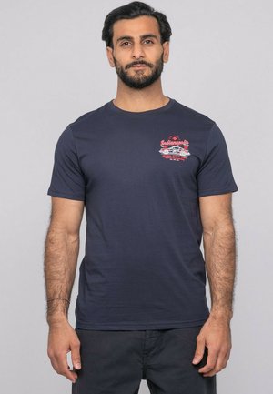Homme barbu portant un t-shirt bleu marine avec un graphisme de voiture vintage rouge et blanche et le texte "Indianapolis" sur la poitrine.