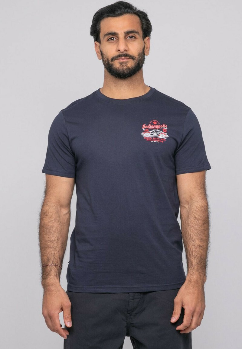 Homme barbu portant un t-shirt bleu marine avec un graphisme de voiture vintage rouge et blanche et le texte "Indianapolis" sur la poitrine.