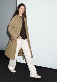 Femme marchant vêtue d'un trench-coat beige, d'un top noir, de pantalons blancs à jambes larges et de ballerines blanches sur un fond noir et blanc.