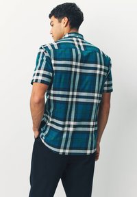 Mann steht mit dem Rücken zur Kamera, trägt ein kurzärmliges Hemd mit blau-grünem Karomuster und dunkle Hosen.