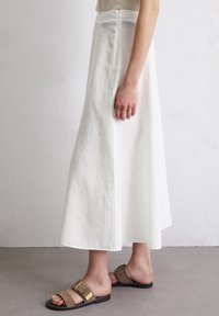 Weißes Leinen-Maxi-Rock mit gerader Silhouette und seitlichem Reißverschluss, kombiniert mit braunen Slide-Sandalen mit goldenen Hardware-Details.