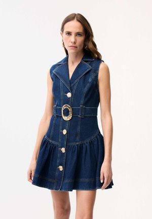 Kobieta w bezrękawowej, ciemnoniebieskiej sukience dżinsowej z dużą złotą klamrą paska i złotymi guzikami, stojąca na białym tle.