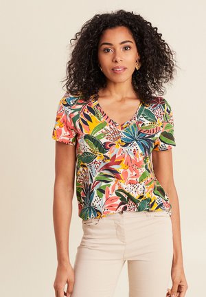 Vrouw met krullend haar, gouden oorbellen en een kleurrijk, kort mouwig V-hals topje met bloemenprint, gecombineerd met lichtbeige broek.