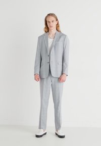 Completo grigio chiaro con giacca monopetto, rever a punta, due tasche frontali e pantaloni abbinati. Indossato con una maglietta bianca e sneaker.