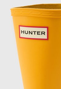 Κίτρινο λαστιχένιο μπότες με υφασμένο επάνω άκρο και ορθογώνια ετικέτα που φέρει το όνομα της μάρκας "HUNTER" με έντονα μαύρα γράμματα σε λευκό φόντο.