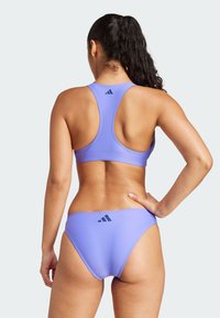 Svijetlo ljubičasta sportska grudnjak i donji dio bikinija s glatkom teksturom, dizajnom s racerback-om i crnim Adidas logotipom na leđima.