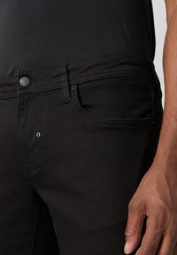 Fekete farmernadrág, slim fit szabással, gombos záródással, elülső zsebekkel és diszkrét varrás részletekkel. Az anyag sima és puha.